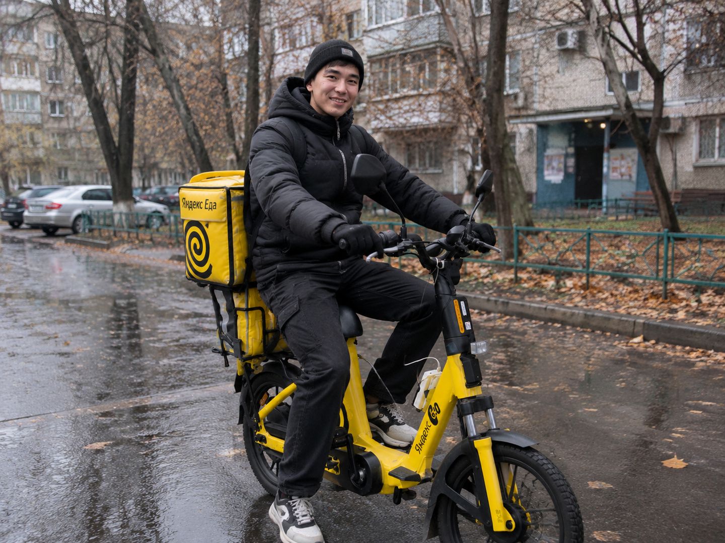 Курьер на жёлтом электровелосипеде с коробом Яндекс Еды во дворе Алматы осенью
