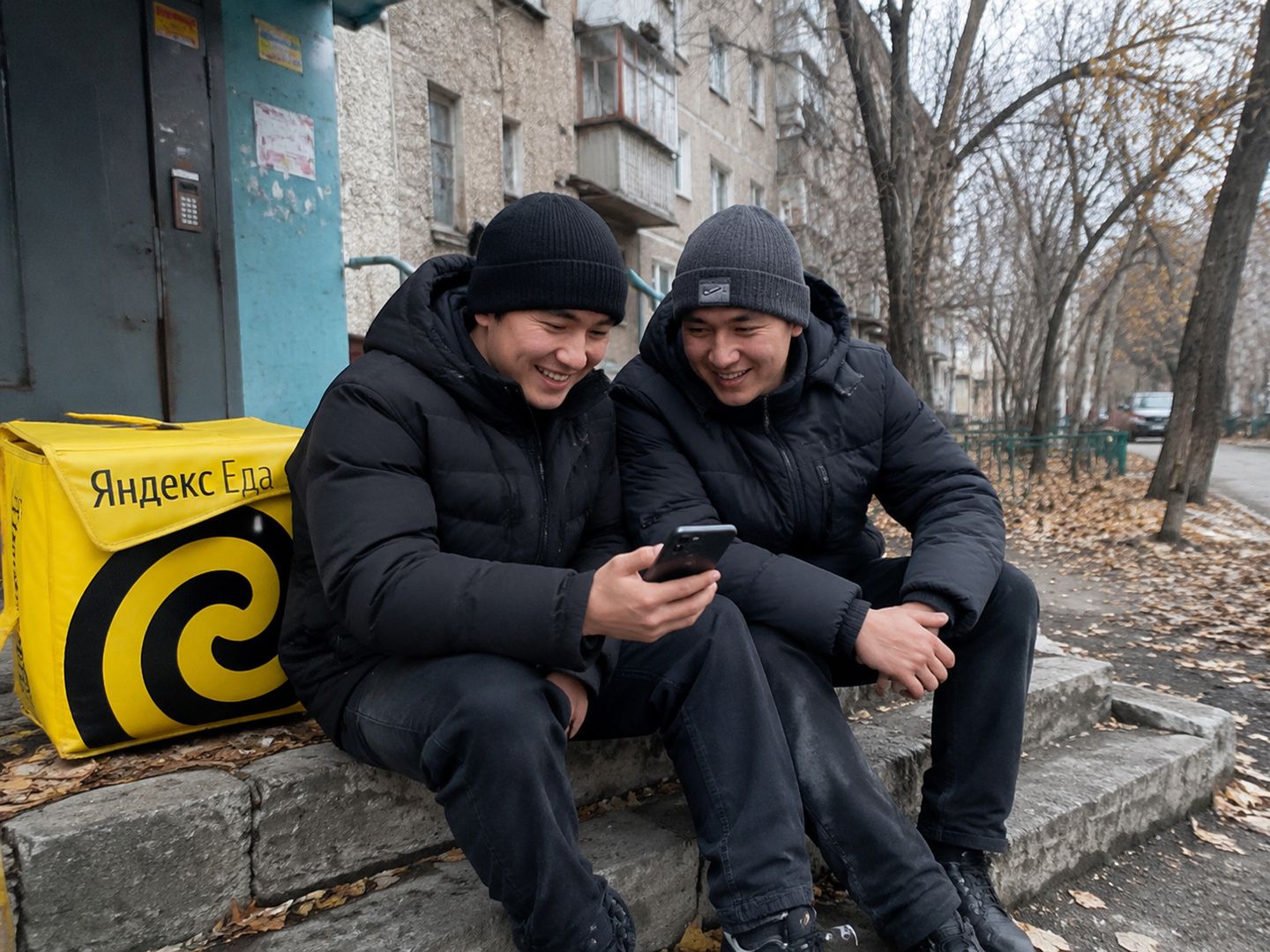 Двое курьеров Яндекс Еды смотрят выплаты в телефоне у подъезда