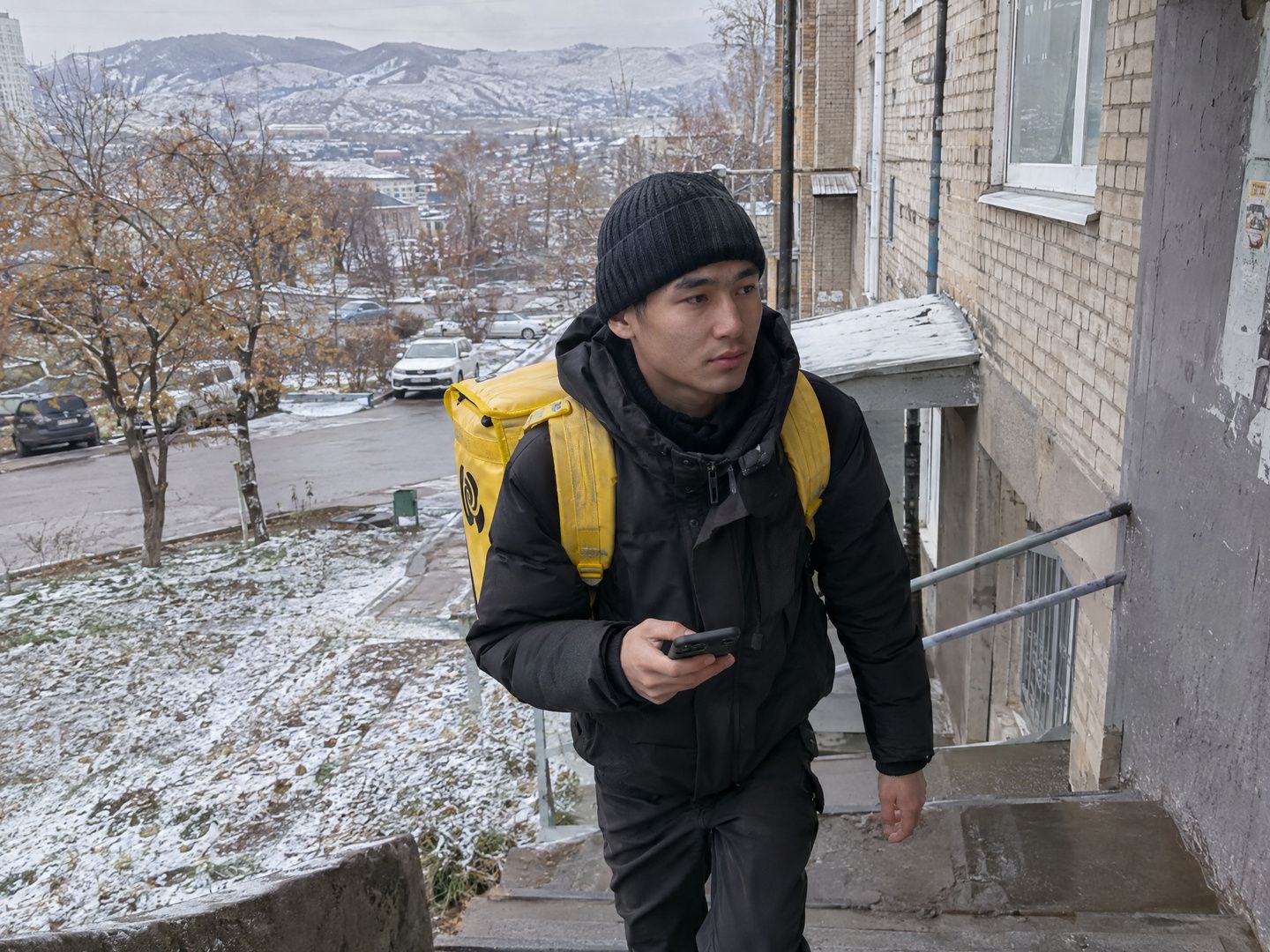 Пеший курьер с жёлтым термокоробом Яндекс Еды поднимается по ступенькам к подъезду в Усть-Каменогорске