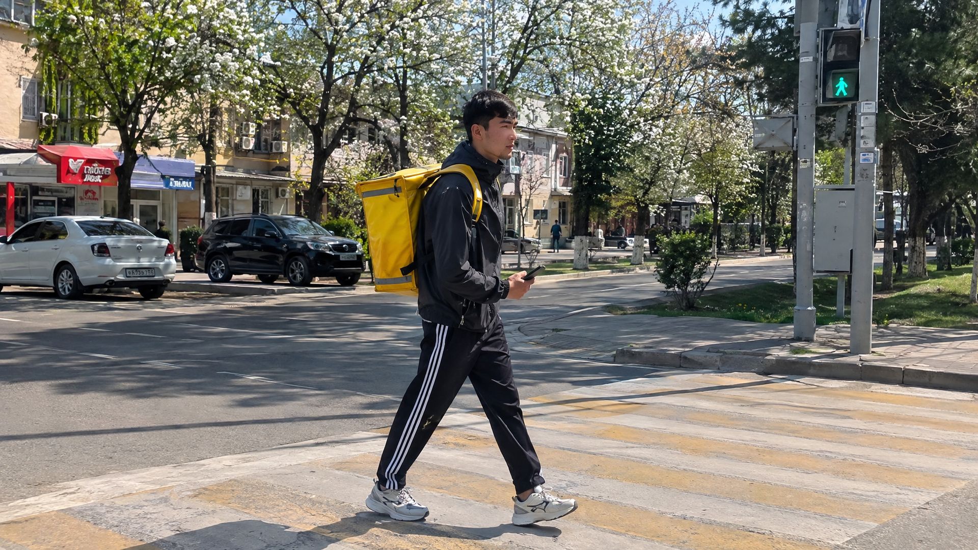 Пеший курьер Яндекс Еды с жёлтым термокоробом переходит улицу по зебре в Шымкенте весной