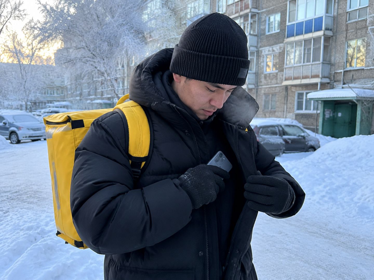 Курьер в тёплых перчатках убирает телефон во внутренний карман на морозе в Усть-Каменогорске